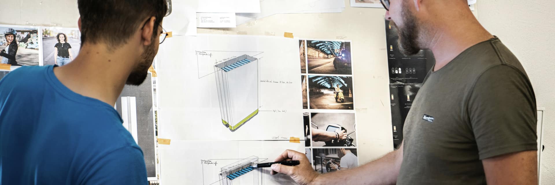 Two engineers reviewing battery design drawings pinned to a wall